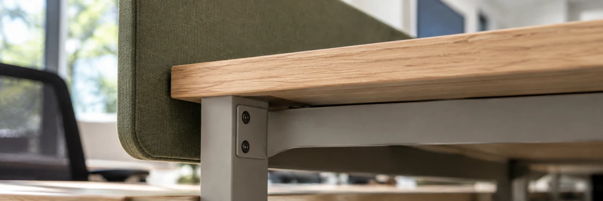 A timber desktop meeting its steel frame at a bolted bracket, a fabric screen behind